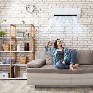 Eine junge Frau sitzt in einem Raum auf der Couch mit der Fernbedienung für die Klimaanlage die an der Wand über Ihr ist. Aus der Klimaanlage kommt Kühle Luft, und die Frau atmet Tief und lächelt.